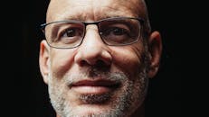 Close-up portrait of a smiling bald elderly man with glasses against a dark background.
